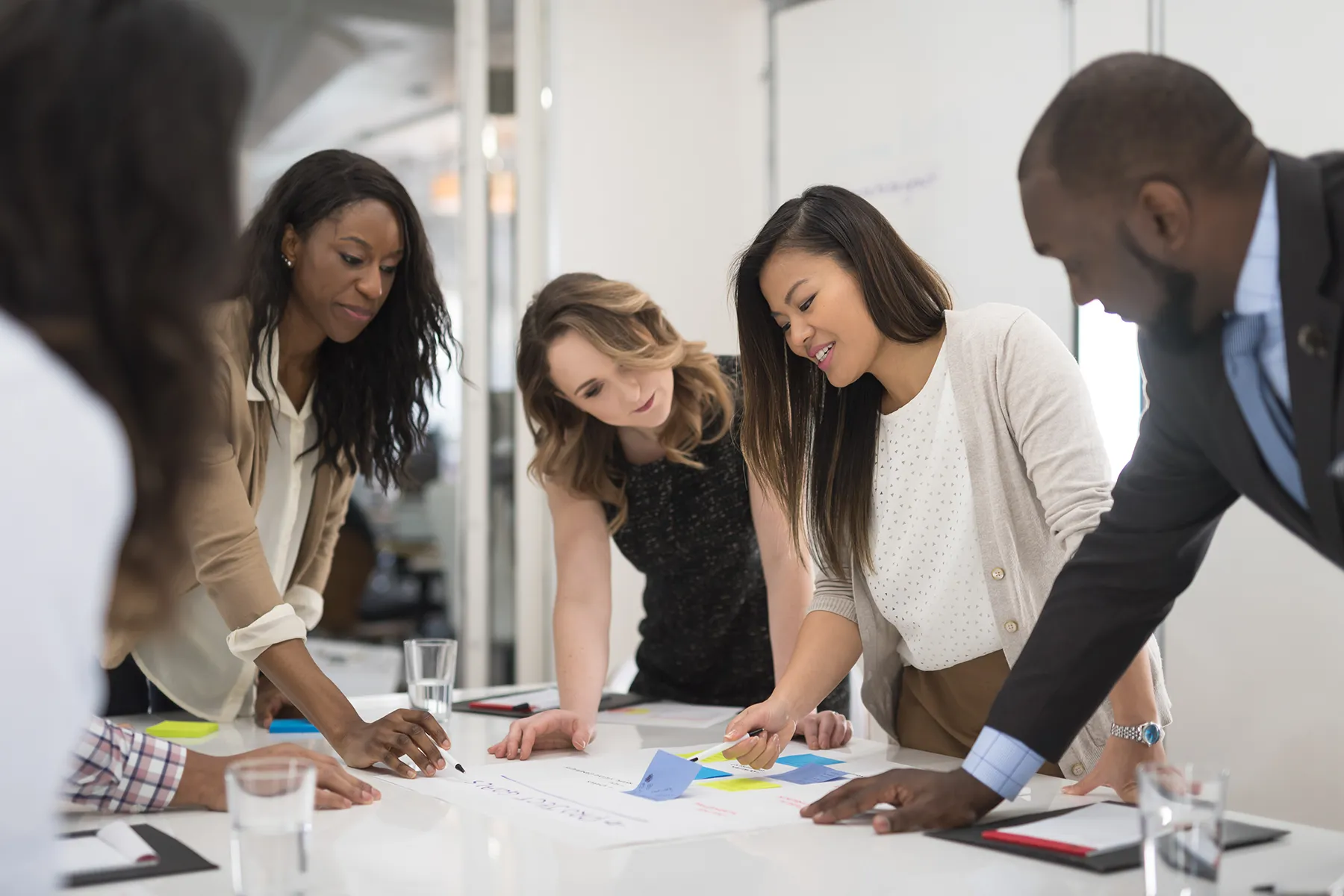 Group of colleagues in business casual attire lean over a white table, examining a schedule covered with colourful sticky notes.