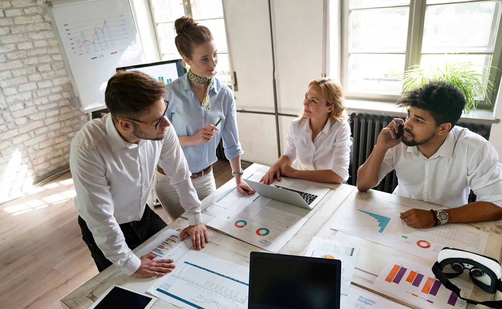 Four people work around a meeting table. There are documents and laptops on the table. One person talks on a cell phone.