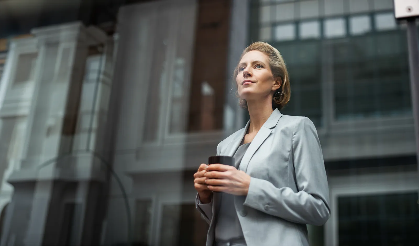 Une femme en tenue d'affaires est debout devant un immeuble avec en main une tasse noire en porcelaine.